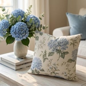 Può includere: Un cuscino quadrato con una stampa botanica di fiori di ortensia blu e foglie verdi su sfondo crema, posizionato accanto a un vaso in ceramica riempito con un bouquet abbinato di ortensie blu fresche e rami di eucalipto.