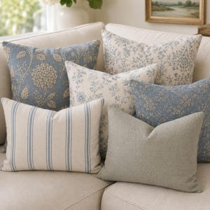 May include: A collection of six decorative throw pillows in various shades of blue, cream, and grey. The set includes square and rectangular cushions featuring floral embroidery, intricate blue and white botanical prints, and blue striped patterns.