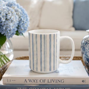 May include: A white ceramic mug featuring a vertical striped pattern in shades of light blue. The mug has a smooth cylindrical body and a curved handle on the right side, positioned upright on top of a stack of two hardcover books.