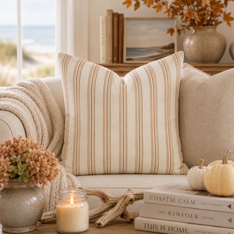 May include: A square throw pillow featuring a vertical striped pattern in shades of cream, burnt orange, and dark blue. The pillow has a smooth, woven texture and is centered, showcasing the repeating linear design across the entire front surface.
