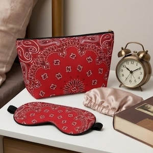 May include: A red bandana-patterned cosmetic bag, sleep mask, and a satin sleep cap are displayed on a white surface. A vintage brass alarm clock and a closed book are also present. The sleep mask has black elastic straps.