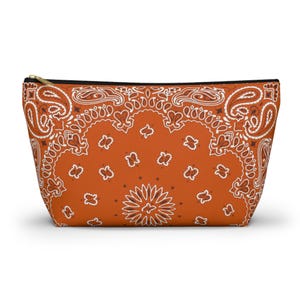 May include: A zippered pouch with a bandana pattern. The pouch is orange with a white paisley design. The zipper is black with a gold-colored pull.