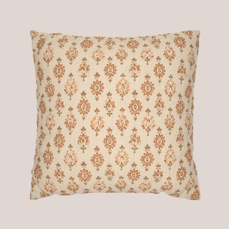 May include: A square throw pillow featuring a cream-colored fabric base with a repeating pattern of small, symmetrical floral motifs in shades of burnt orange and muted brown, arranged in vertical columns across the entire surface of the cushion.