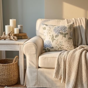 May include: A cream-colored sofa features a square throw pillow with a blue hydrangea and botanical print. A beige knit throw blanket is draped over the arm. Beside the sofa, a white wooden side table holds three pillar candles and books.