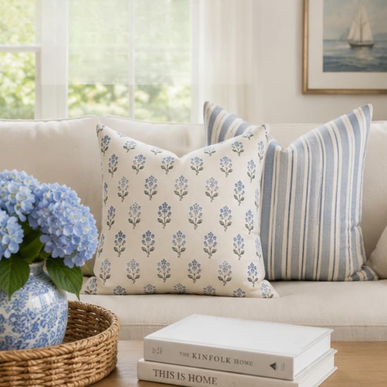 May include: Two square throw pillows on a sofa. One pillow features a repeating blue floral pattern on an off-white background. The second pillow has vertical blue and white stripes. A blue and white vase with blue hydrangeas sits nearby.
