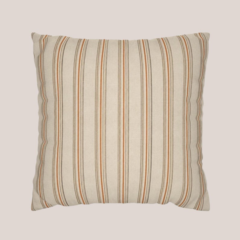 May include: A square throw pillow featuring a vertical striped pattern in shades of burnt orange, dark charcoal, and light gray against a neutral beige background. The fabric has a visible woven texture, and the stripes repeat across the surface.