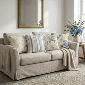 May include: A beige sofa with a slipcover, draped with a textured throw blanket, featuring four decorative pillows: two with blue hydrangea floral patterns, one with a blue striped pattern, and one with a delicate botanical wildflower print.