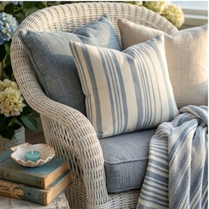 May include: A white wicker armchair featuring a solid blue cushion, a blue textured throw pillow, a blue and white striped throw pillow, a beige throw pillow, and a blue and white striped throw blanket draped over the seat.