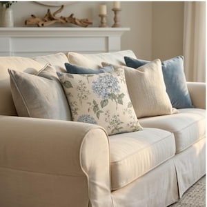 May include: A cream-colored sofa featuring a collection of throw pillows in various textures and colors, including a central pillow with a blue hydrangea floral pattern, solid light blue velvet pillows, and plain beige linen-style cushions.