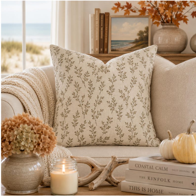 May include: A square throw pillow featuring a light beige fabric base with a repeating pattern of small, dark brown botanical leaf sprigs. The pillow has a structured, plump appearance and is positioned centrally, showcasing a clean, woven texture.