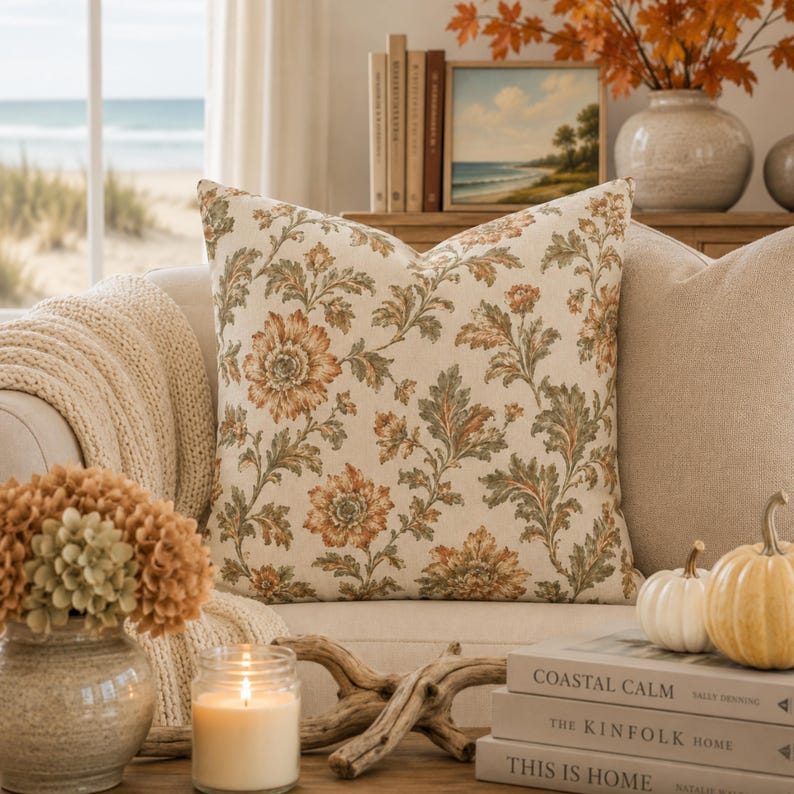 May include: A square throw cushion featuring a cream-coloured fabric base with a repeating botanical pattern of orange-brown flowers and sage green leaves. The cushion is displayed on a sofa, showcasing a detailed floral print across the front surface.