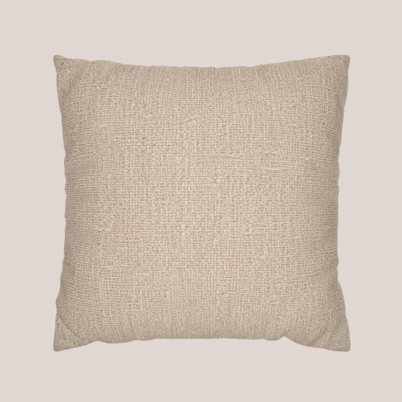 May include: A square throw pillow featuring a solid light beige color and a prominent, coarse woven fabric texture. The pillow has a simple, clean-edged design with a visible grid-like weave pattern throughout the entire surface of the cover.
