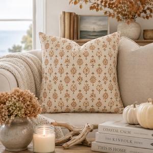 May include: A square throw pillow featuring a repeating damask-style pattern in shades of burnt orange, tan, and brown on a cream-colored fabric base. The pillow has a structured, plump appearance with clean, finished edges and a centered design.