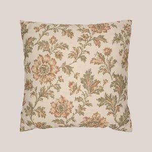May include: A square throw pillow featuring a cream-colored base fabric with a repeating floral pattern. The design consists of large, detailed flowers in shades of muted orange and brown, accented by intricate green foliage and leafy stems.