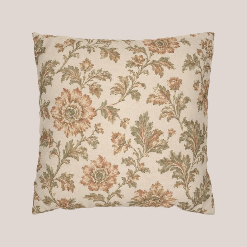 May include: A square throw pillow featuring a cream-colored base fabric with a repeating floral pattern. The design consists of large, detailed flowers in shades of muted orange and brown, accented by intricate green foliage and leafy stems.