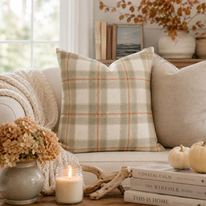 May include: A square throw pillow featuring a plaid pattern in shades of cream, sage green, and thin orange lines. The fabric has a visible woven texture. The pillow is positioned upright, displaying a symmetrical grid design across its surface.