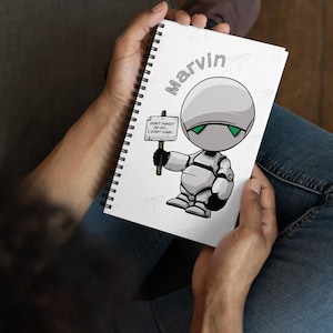 May include: A spiral-bound notebook with a cartoon robot illustration. The robot is gray and white, with green eyes, and holds a sign that reads "DON'T PANIC! OR DO... I DON'T CARE...". The name "Marvin" is above the robot.