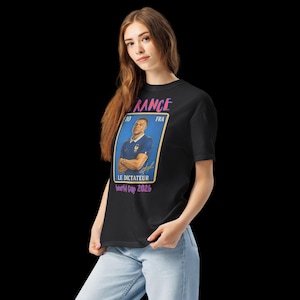 May include: Black t-shirt featuring a graphic of a soccer player in a blue jersey. The graphic includes the word "FRANCE" in pink, the text "LE DICTATEUR" and "World Cup 2026". The shirt is worn by a person.