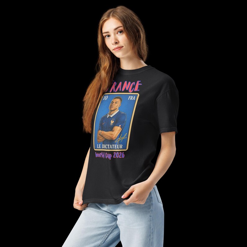 May include: Black t-shirt featuring a graphic of a soccer player in a blue jersey. The graphic includes the word "FRANCE" in pink, the text "LE DICTATEUR" and "World Cup 2026". The shirt is worn by a person.