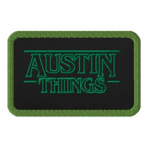 May include: Embroidered patch with the words "AUSTIN THINGS" in green thread on a black background. The patch has a green border. The text is in a stylized font.