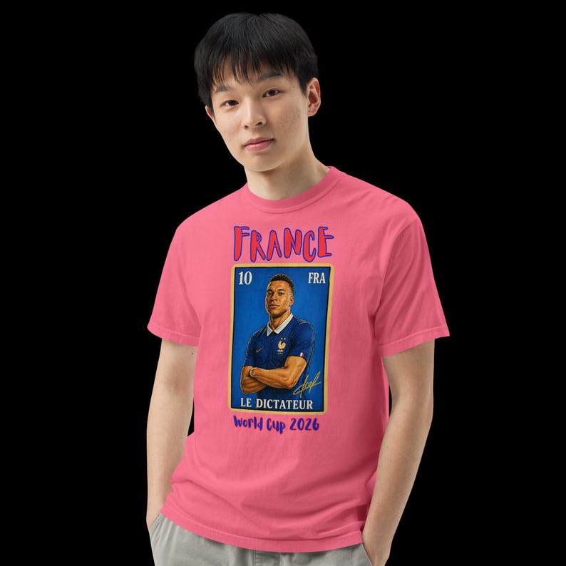 May include: A pink t-shirt featuring a graphic of a soccer player in a blue jersey. The graphic includes the word "FRANCE" in purple, the number "10", and the text "World Cup 2026". The shirt is worn by a person.