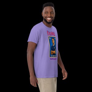 May include: A light purple t-shirt featuring a graphic of a French soccer player in a blue jersey. The graphic is designed to look like a postage stamp with the word "FRANCE" at the top. The text "LE DICTEUR World Cup" is at the bottom.