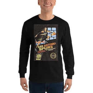 May include: Black long-sleeve shirt featuring a graphic of a basketball player in action, with the text "San Antonio Spurs Entertainment System." The design includes the team's logo and a vintage aesthetic.