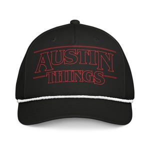 Könnte beinhalten: Schwarze Baseballkappe mit dem roten Text "AUSTIN THINGS" und einem weißen Seildetail am Rand. Die Kappe hat einen gebogenen Schirm und einen Knopf oben.