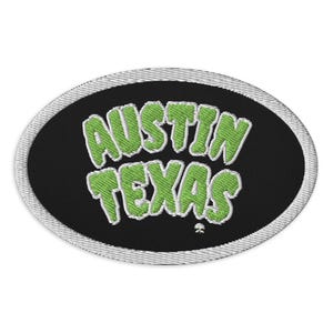 May include: An embroidered patch featuring the text "AUSTIN TEXAS" in green with a white outline, set against a black oval. The patch has a white border.