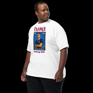 May include: White t-shirt featuring a portrait of a soccer player in a blue jersey. The shirt has the word "FRANCE" in red and blue, along with the text "LE DICTATEUR World Cup 2026". The shirt is worn by a person.