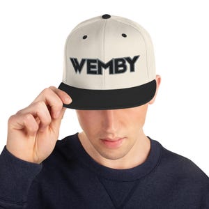 Pode incluir: Boné de beisebol creme com aba preta. A palavra "WEMBY" é bordada em preto e cinza na frente do boné. O boné está sendo segurado por uma pessoa.