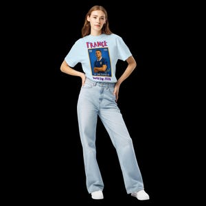 May include: A light blue t-shirt with a graphic of a postage stamp featuring a soccer player in a blue jersey. The stamp reads "FRANCE" and "LE DICTATEUR World Cup 2026". The person is wearing light blue jeans and white sneakers.