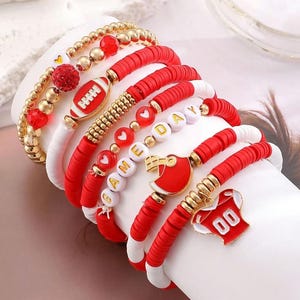 May include: Stack of red, white, and gold beaded bracelets with football and jersey charms. Some bracelets have the words "GAME DAY" spelled out with white letter beads. The bracelets are on a white surface.
