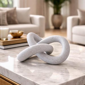 May include: A light gray decorative knot sculpture made of stone sits on a marble-topped coffee table with a gold-colored wooden frame. The sculpture has a speckled texture and a looped design. The background includes a living room setting with white sofas and a large potted plant.
