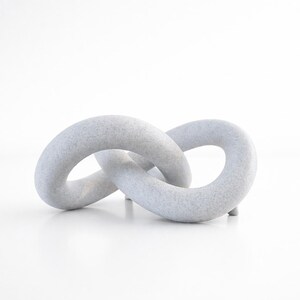May include: A light grey, abstract sculpture in the shape of two interlocking loops. The sculpture has a textured surface and sits on a white surface. The piece is a decorative object.