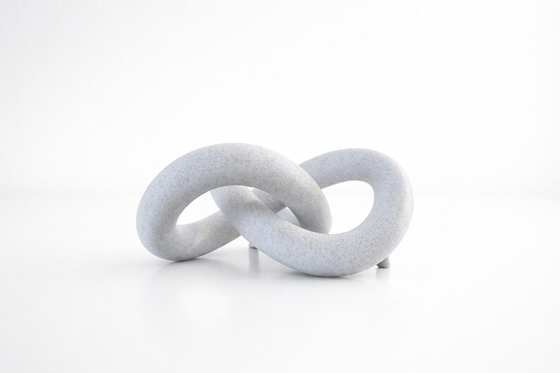 May include: A light grey, abstract sculpture in the shape of two interlocking loops. The sculpture has a textured surface and sits on a white surface. The piece is a decorative object.