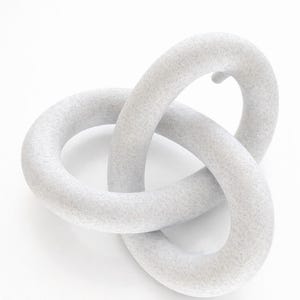 May include: A light grey, abstract sculpture with a textured surface. The sculpture is composed of two interlocking, rounded forms, creating a continuous loop. The piece is set against a white background.