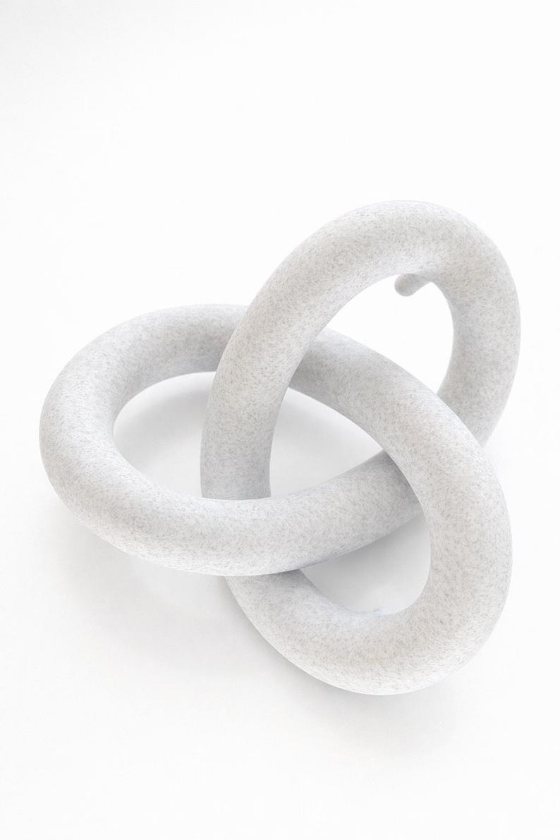 May include: A light grey, abstract sculpture with a textured surface. The sculpture is composed of two interlocking, rounded forms, creating a continuous loop. The piece is set against a white background.