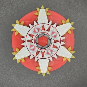 May include: A 3D-printed, multi-layered object with a red base, a white and red central design, and yellow accents. The design features a circular shape with a complex, geometric pattern, resembling a stylized flower or gear.