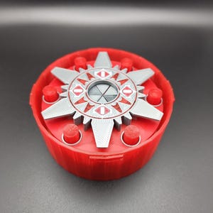 May include: A red and silver 3D-printed puzzle box. The box has a circular base with a star-shaped top piece. The top piece has a series of eye-shaped designs and is surrounded by red cylinders.