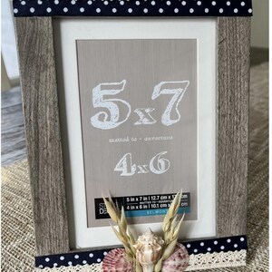 May include: A decorative wooden picture frame with a grey finish, accented with navy blue polka dot ribbon and cream lace. The frame is embellished with seashells and wheat. The frame is designed for 12.7cm x 17cm and 10.1cm x 15.2cm photos.