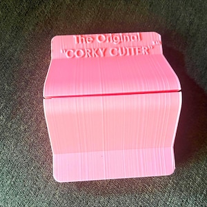 May include: A pink "CORKY CUTTER" wine cork remover. The text "The Original" is above the product name. The tool has a rectangular shape with a curved top and a textured surface.