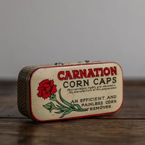 Op de afbeelding: Vintage Carnation Corn Caps blik. Het rechthoekige blik heeft een crèmekleurige achtergrond met rode tekst en een rode bloemillustratie. De tekst luidt "Carnation Corn Caps" en "Een efficiënte en pijnloze eeltverwijderaar."