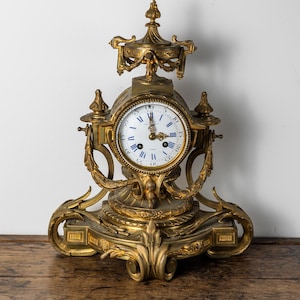 May include: Ornate gold-colored mantel clock with a white clock face and blue Roman numerals. The clock features intricate detailing, including decorative flourishes and a top finial. The clock is displayed on a wooden surface.
