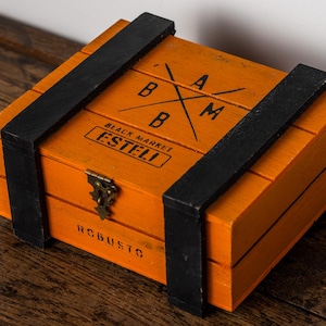 May include: Orange wooden box with black accents and a metal clasp. The box has the text "BAM" and "BLACK MARKET ESTELI" printed on the top. Black straps secure the lid. The word "ROBUSTO" is printed on the front.