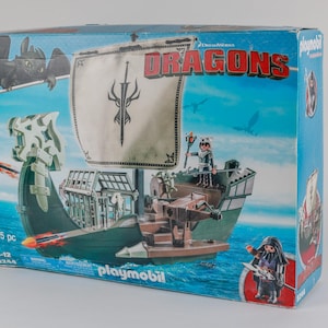 Puede incluir: Caja de Playmobil con un barco de la película "Cómo entrenar a tu dragón". La caja azul muestra imágenes del barco, personajes y un dragón. El barco tiene una vela blanca con un emblema negro sobre un fondo de océano azul.