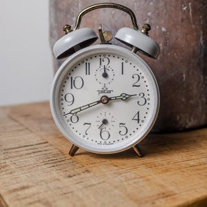 Könnte beinhalten: Ein weißer Vintage-Wecker mit cremefarbenem Zifferblatt, schwarzen Ziffern und Zeigern. Die Uhr hat einen goldfarbenen Griff und Glocken und steht auf einer Holzoberfläche. Auf dem Zifferblatt steht "peter".