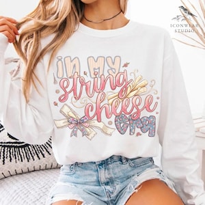 May include: White long-sleeve sweatshirt featuring the text "In My String Cheese Era" in pink and blue script. The design includes illustrations of string cheese, a bow, and floral accents. The sweatshirt is a casual, comfortable style.