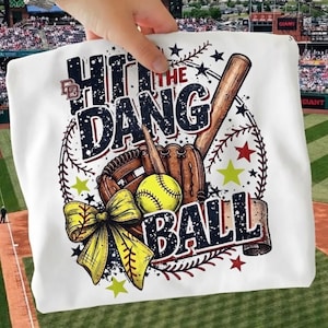 May include: White t-shirt with a baseball-themed graphic. The design includes a baseball bat, glove, and ball, with the text "Hit the Dang Ball." The design also features a yellow bow and stars.