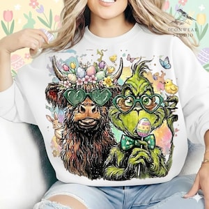 May include: White sweatshirt featuring a whimsical Easter design. The graphic includes a brown Highland cow and a green Grinch character, both wearing heart-shaped sunglasses and Easter-themed accessories. The design is surrounded by pastel colors and Easter elements.